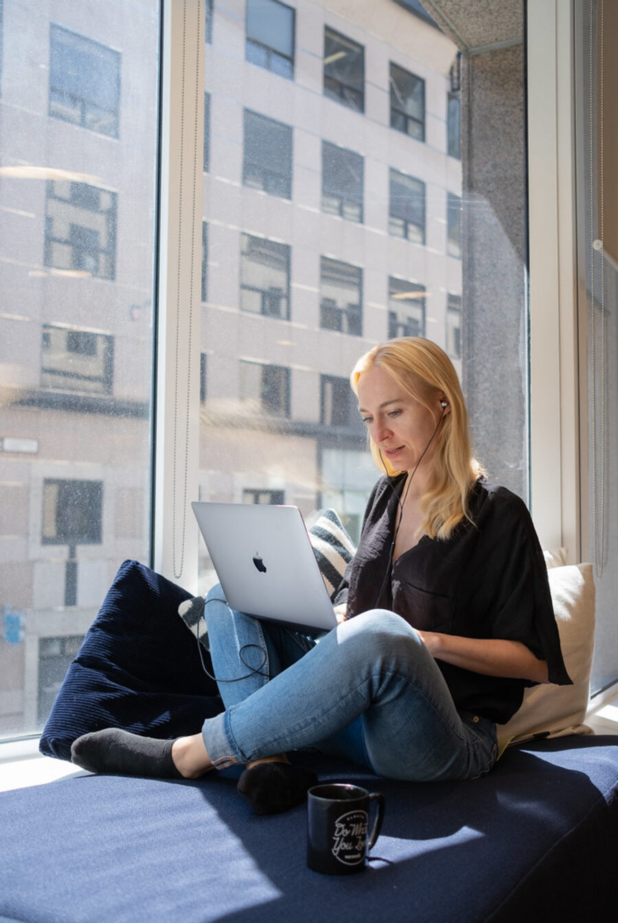 A woman sitting by a window, listening on earbuds while reading on her laptop.